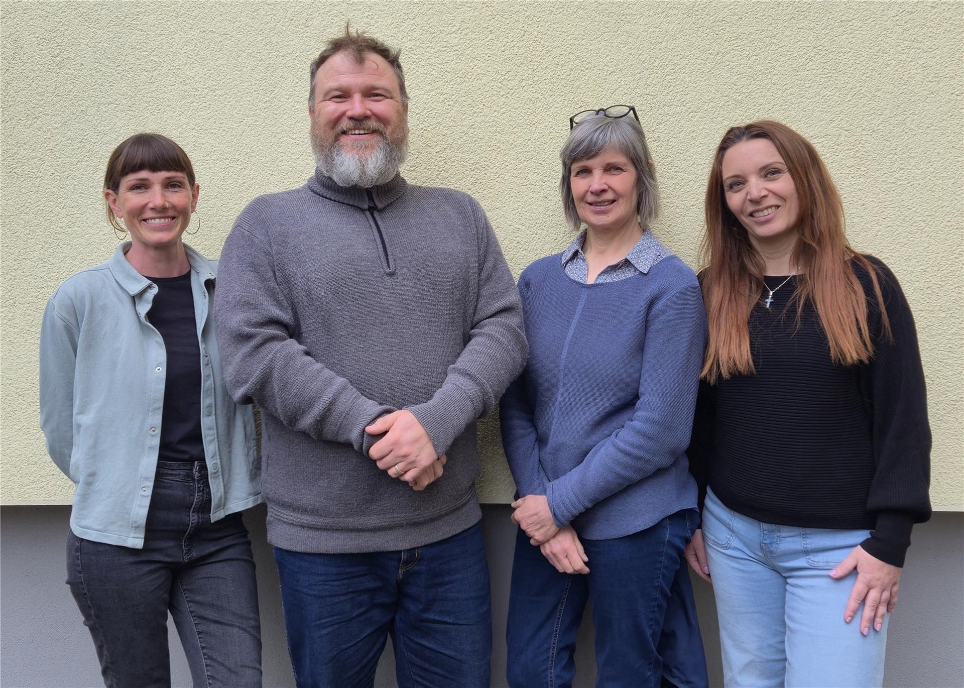 Das Bild zeigt die vier Mitarbeitenden der Organisierten Nachbarschaftshilfen in Nahansicht vor einer hellen Mauer des Caritas-Gebäudes in Ravensburg. 